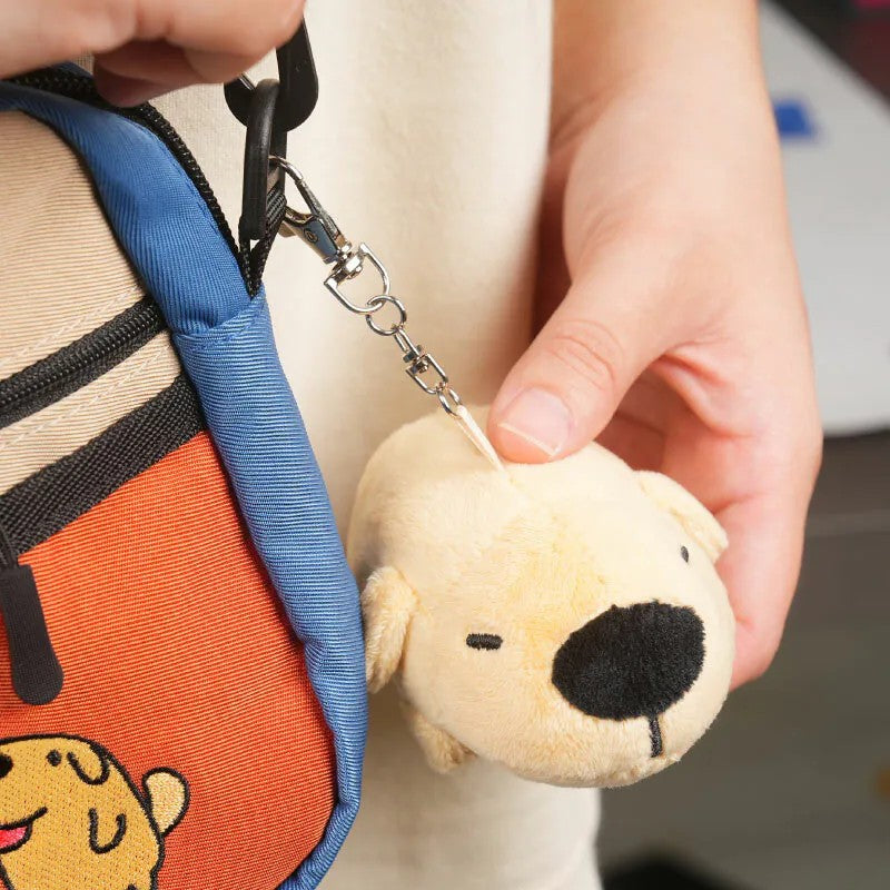 Hand holding the Dogdog Lazy Plush Charm, showing it being clipped onto a bag with a metal chain.